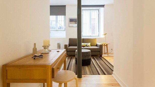 Een moderne woonkamer met een bank, een salontafel en een eettafel met stoelen. Er is een raam met zonwering, een lamp en een schilderij aan de muur.