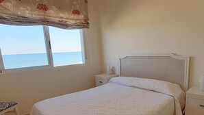 Un dormitorio con cama, mesita de noche y cortina, con ventana y vista al mar.