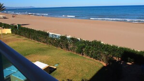 Vista a la playa con cielo despejado, arena y una zona con césped y una valla.