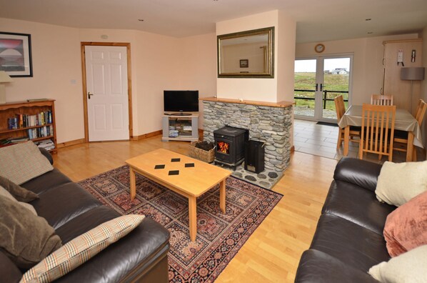 A living room with a fireplace, a television, a bookshelf, and a dining area with chairs and a table. - Cottage 102 - Ballyconneely (Ballyconneely)