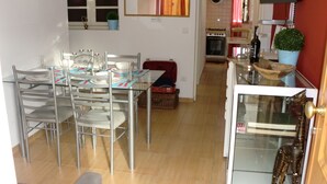 Un coin repas avec une table en verre et des chaises en métal, une valise rouge posée sur le sol, et une cuisine en arrière-plan.