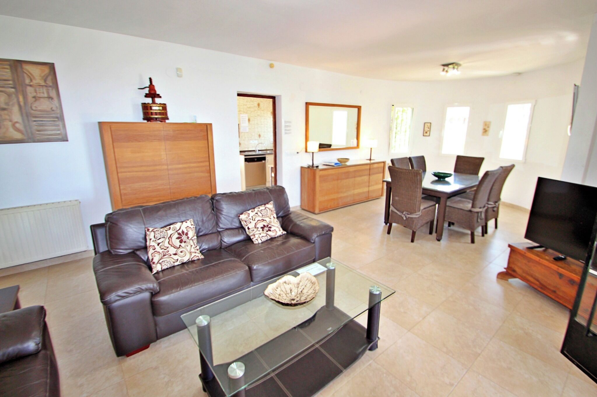 A modern living room with a brown leather sofa, a glass coffee table, and a dining area with a wooden table and chairs.