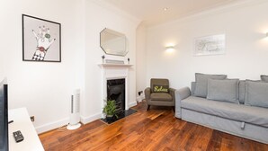 Een woonkamer met een open haard, een grijze bank, een fauteuil met een 'Relax' kussen en een gekaderd kunstwerk aan de muur.