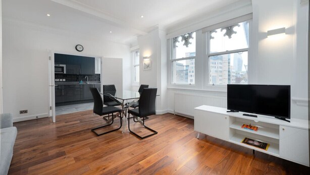 Een moderne woonkamer met een eettafel, stoelen, een flatscreen-tv en een keuken op de achtergrond.