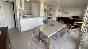 Eine moderne Küche mit weißen Schränken, ein Essbereich mit einem weißen Tisch und transparenten Stühlen sowie ein schwarzes Sofa im Wohnbereich.