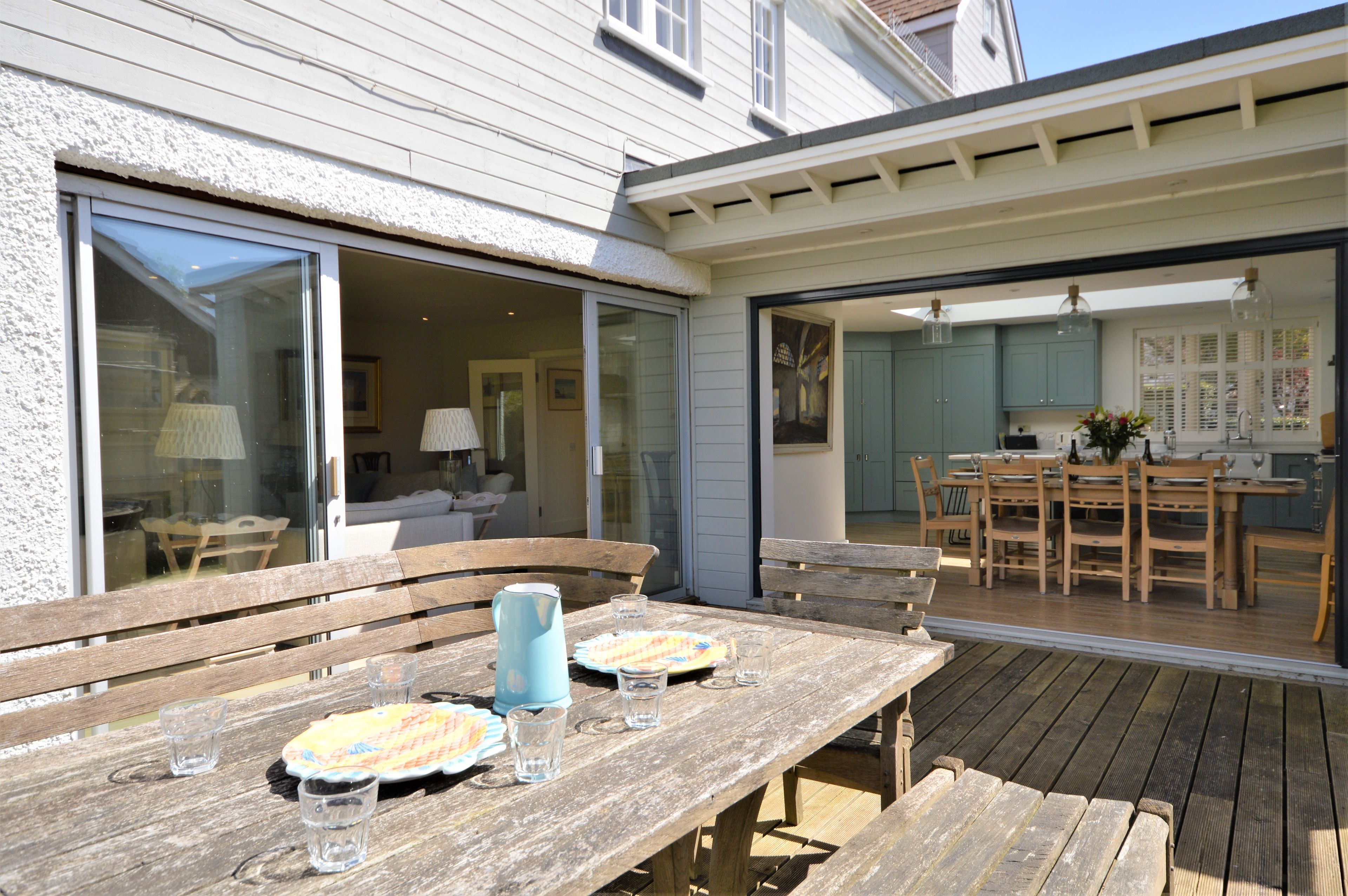 An outdoor dining area with wooden tables and benches, a kitchen visible through an open door, and a patio with a wooden deck.