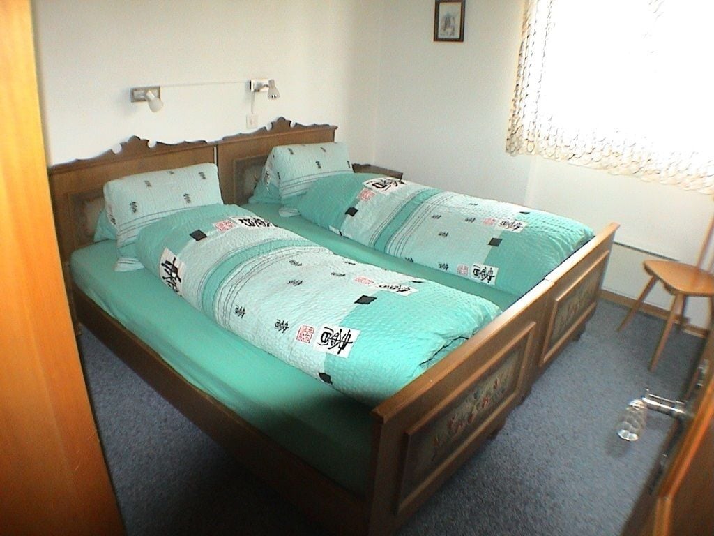 A double bed with a wooden frame and green bedding. There is a small wooden stool and a wall-mounted light fixture.