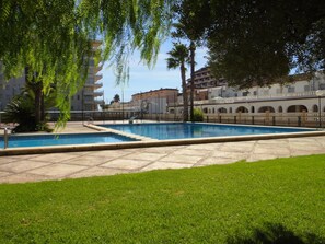 Relax by the serene pool surrounded by lush greenery and palm trees, overlooking a classic building with balconies and arched windows.