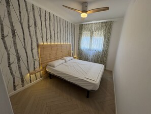 Un dormitorio con cabecera de madera, cama con ropa de cama blanca, mesita de noche, ventilador de techo y ventana con cortinas.
