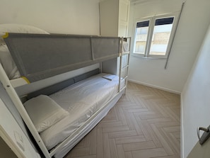 Una cama litera con cabecera gris, ropa de cama blanca y una ventana con persianas.