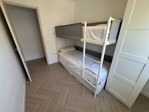 Une chambre avec un lit superposé, une porte, un parquet à motifs en chevrons et une armoire blanche.