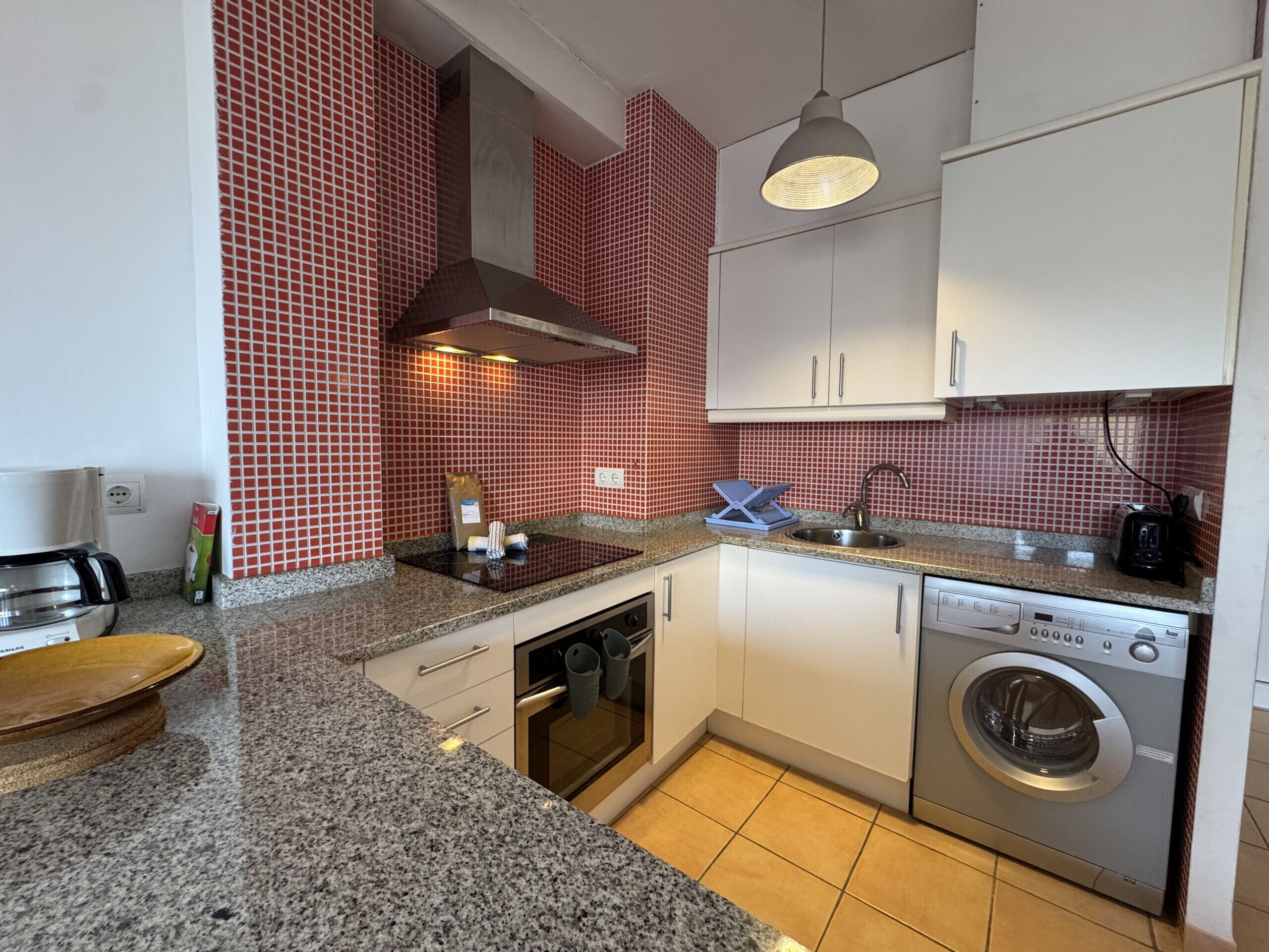 Een keuken met een granieten aanrecht, een ingebouwde oven, een wasmachine en een rode tegelachtergrond.