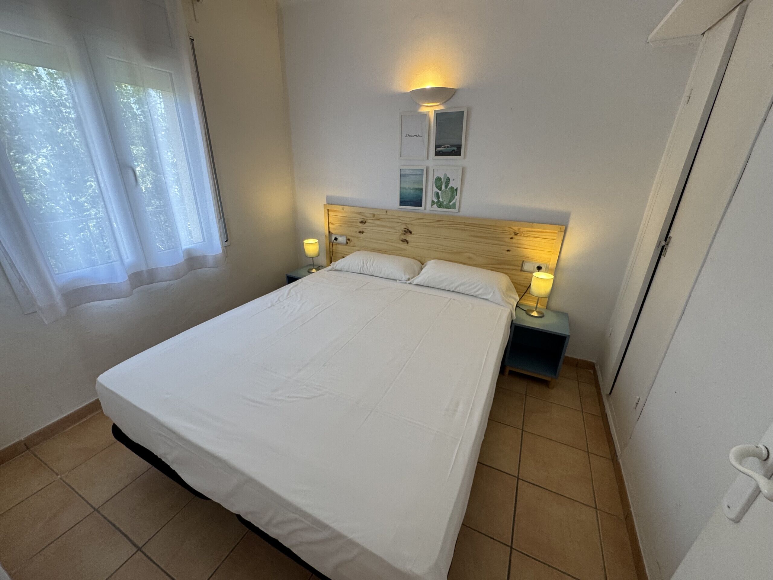 A neatly made bed in a room with a wooden headboard, two bedside tables with lamps, a window with sheer curtains, and a wall-mounted light fixture.