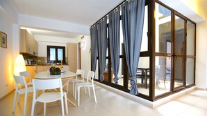 A modern kitchen with white cabinets, a dining area with a round table and white chairs, and a large sliding glass door with blue curtains.