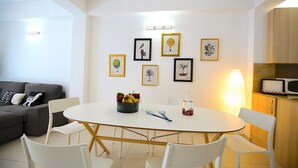 A modern living room with a round white table, white chairs, a grey sofa, and a kitchen area with a microwave.