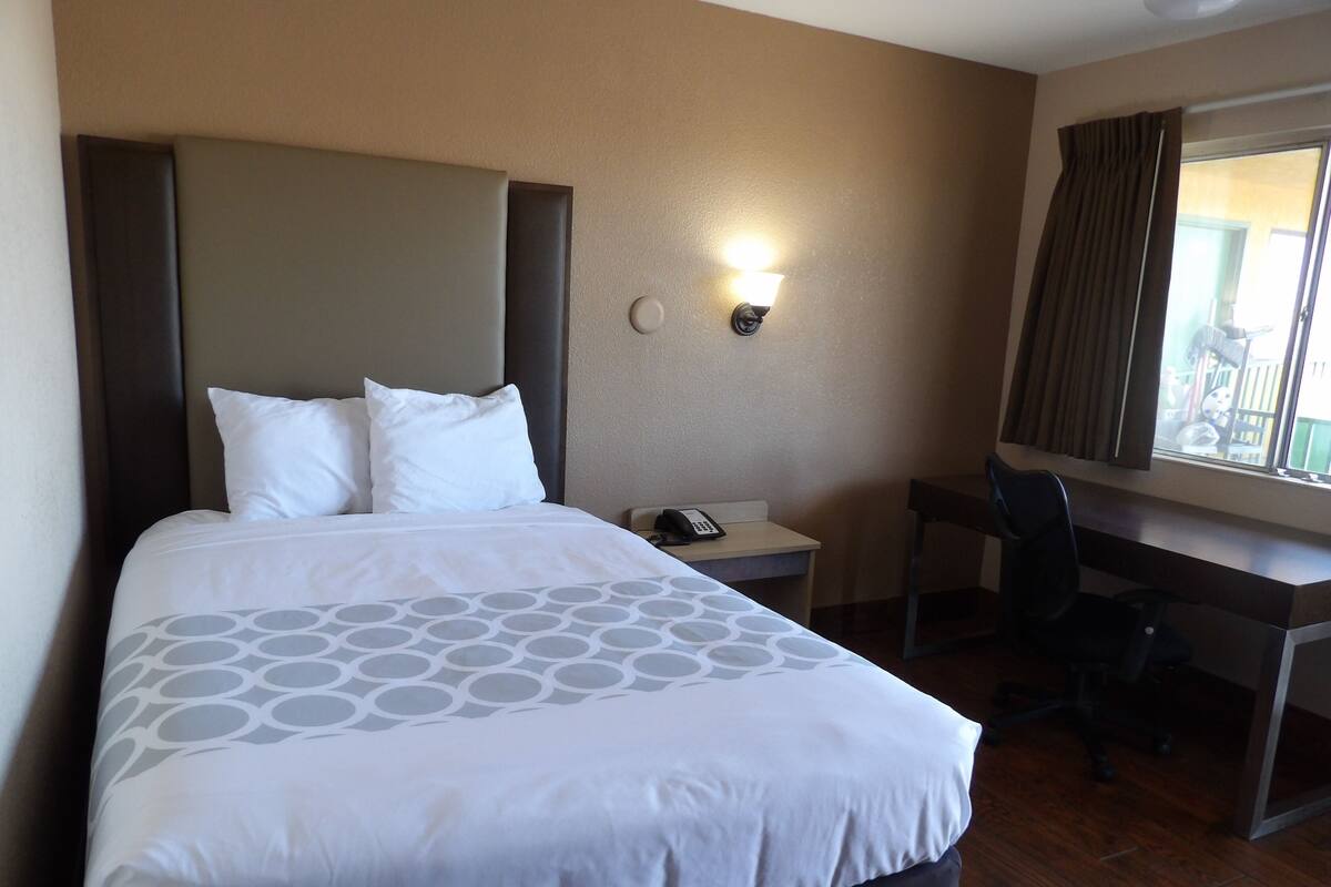Une chambre d’hôtel avec un lit, un bureau, une chaise, un téléphone et une fenêtre avec des rideaux.