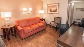 A hotel room with a wooden floor, an orange sofa, a wooden table, two lamps, a framed picture, and a chair.