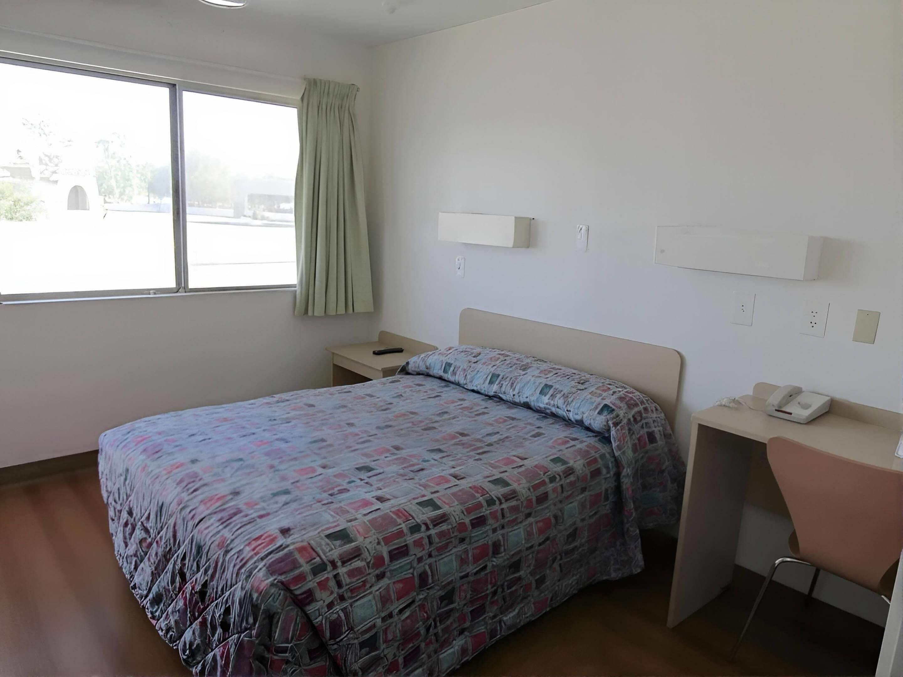 A hotel room with a bed, a desk, a chair, a window with curtains, and a view of the outside.