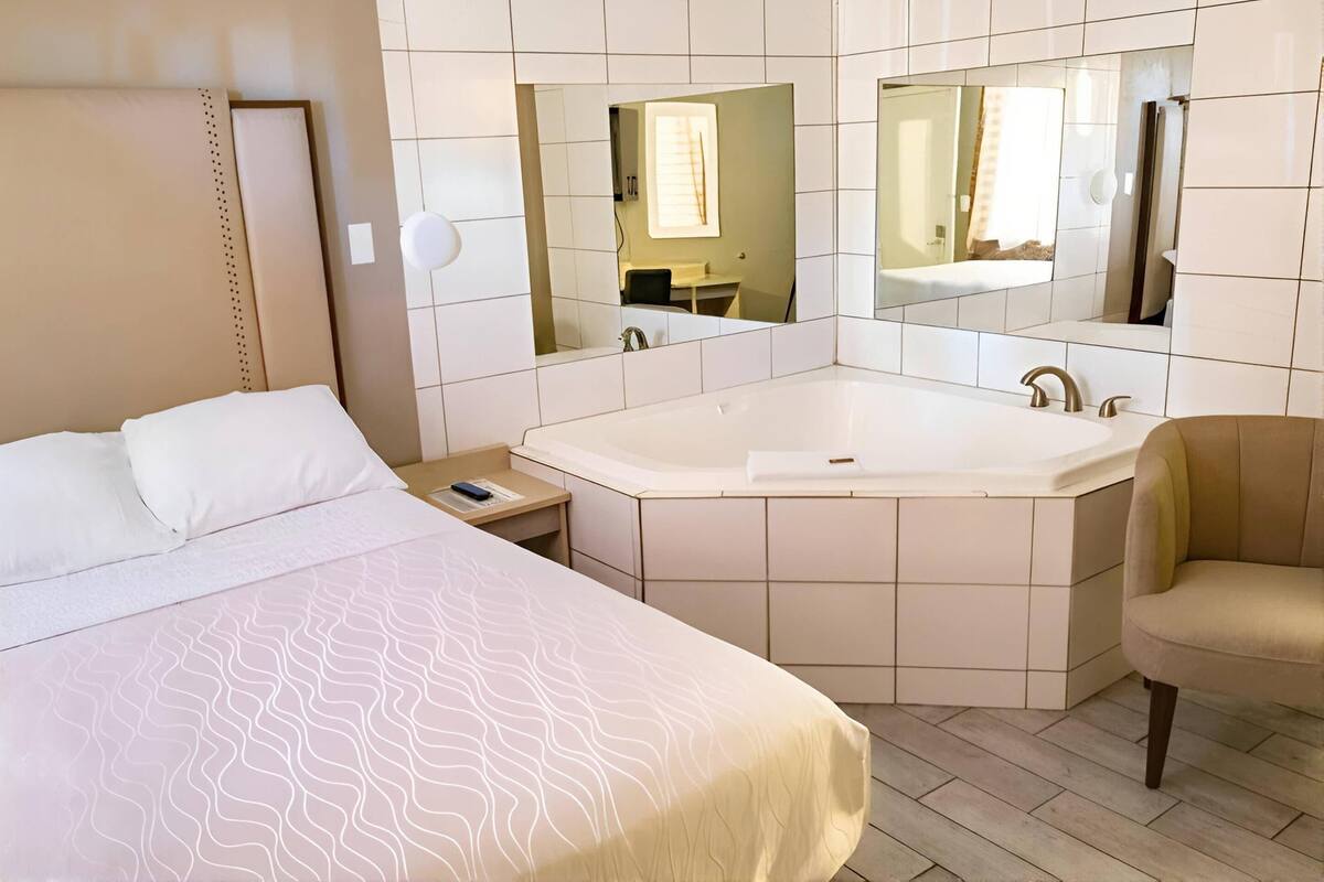 Une chambre d’hôtel avec un lit, une grande baignoire, une chaise, une table de chevet et un miroir.