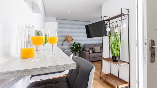 A modern living room with a dining area, a flat-screen TV mounted on the wall, and a wooden shelf with plants. - San Francisco Penthouse Studio (Sevilla)