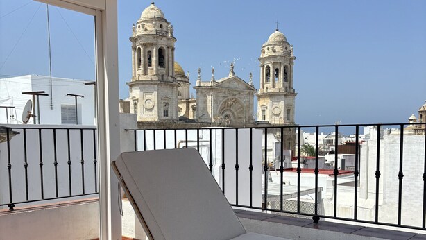 A balcony with a white cushioned lounge chair and a wooden table, overlooking a historic building with domes and bell towers.
