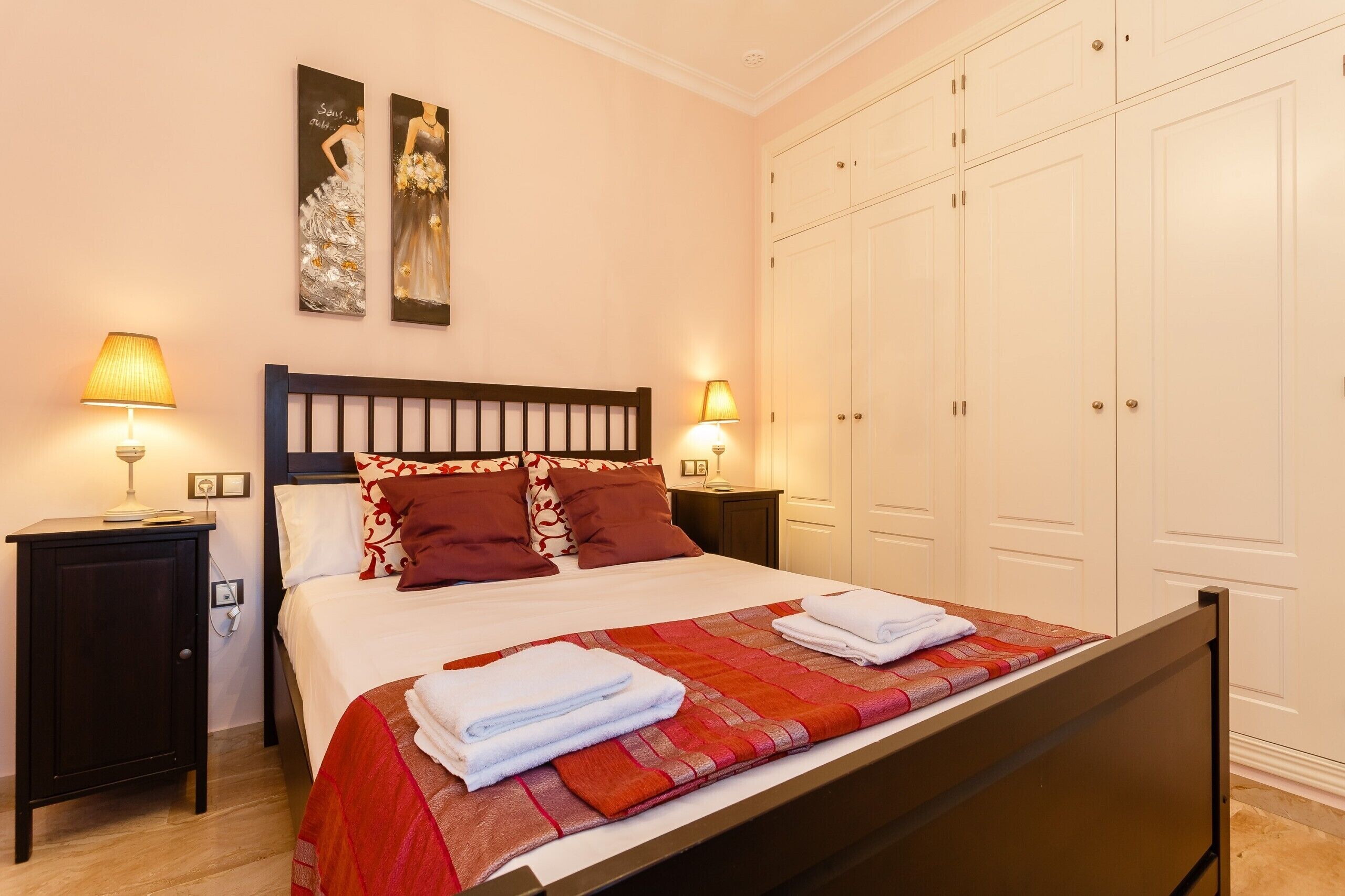 A neatly made bed with a red and orange blanket, two bedside lamps, and a wardrobe with multiple doors.