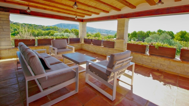 Eine Terrasse im Freien mit weißer Möbel, Steinmauern und Blick auf Grünflächen und Hügel.