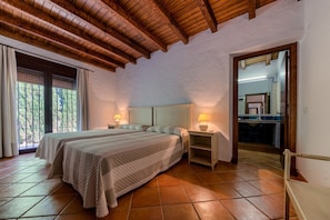A bedroom with a wooden ceiling, a bed with a quilt, a nightstand with a lamp, and a bathroom visible through an open door.