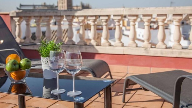 Una mesa con un vaso de agua, dos copas de vino y un bol de fruta en una terraza con vistas a una torre y barandilla.