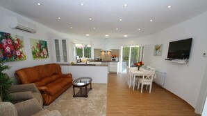 Ein modernes Wohnzimmer mit einer braunen Ledersofa, einem grauen Sessel und einem Glastisch. Ein Essbereich mit weißem Tisch und Stühlen sowie eine Küchenzeile mit weißen Schränken und einem Fenster.