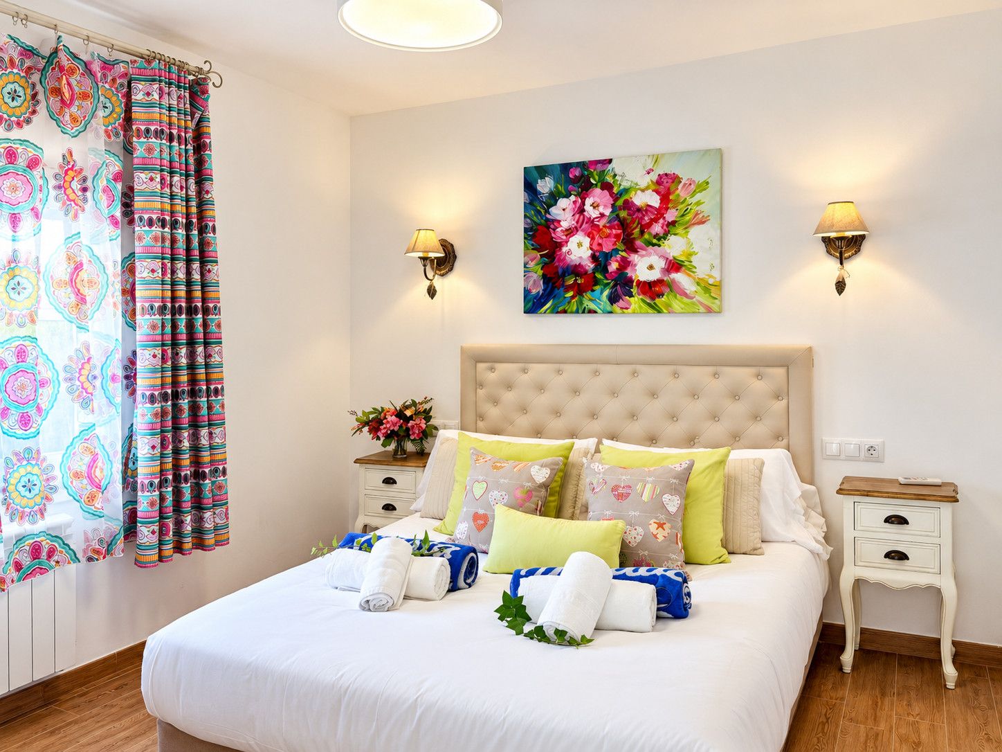 Une chambre d’hôtel avec un lit, deux tables de chevet, une petite table, un tableau représentant des fleurs et un rideau à motifs colorés.