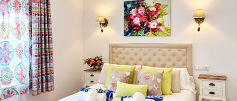 A hotel room with a bed, two bedside tables, a nightstand, a floral painting, and a curtain with a colorful pattern.