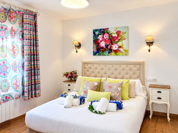 A hotel room with a bed, two bedside tables, a nightstand, a floral painting, and a curtain with a colorful pattern.
