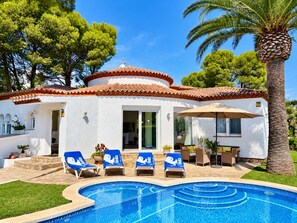 A white building with a tiled roof, a swimming pool, and lounge chairs with umbrellas.