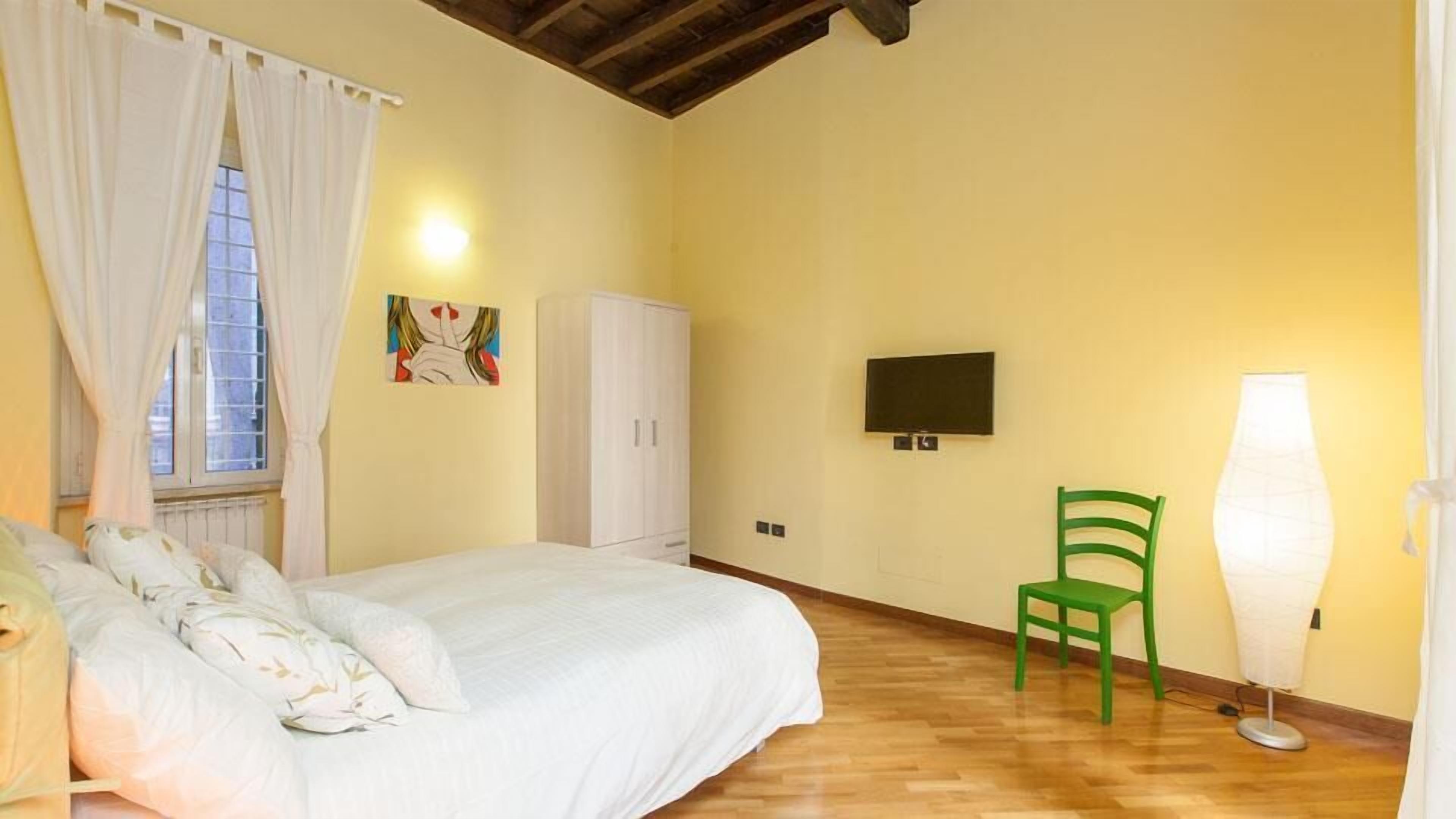 A bedroom with a bed, a green chair, a white wardrobe, a TV, and a floor lamp.