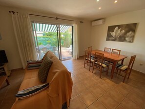 Ein Wohnzimmer mit einer Couch, einem Esstisch mit Stühlen, einem Fernseher und einer Schiebetür zu einem Außenbereich mit Terrasse und Sitzgelegenheiten.