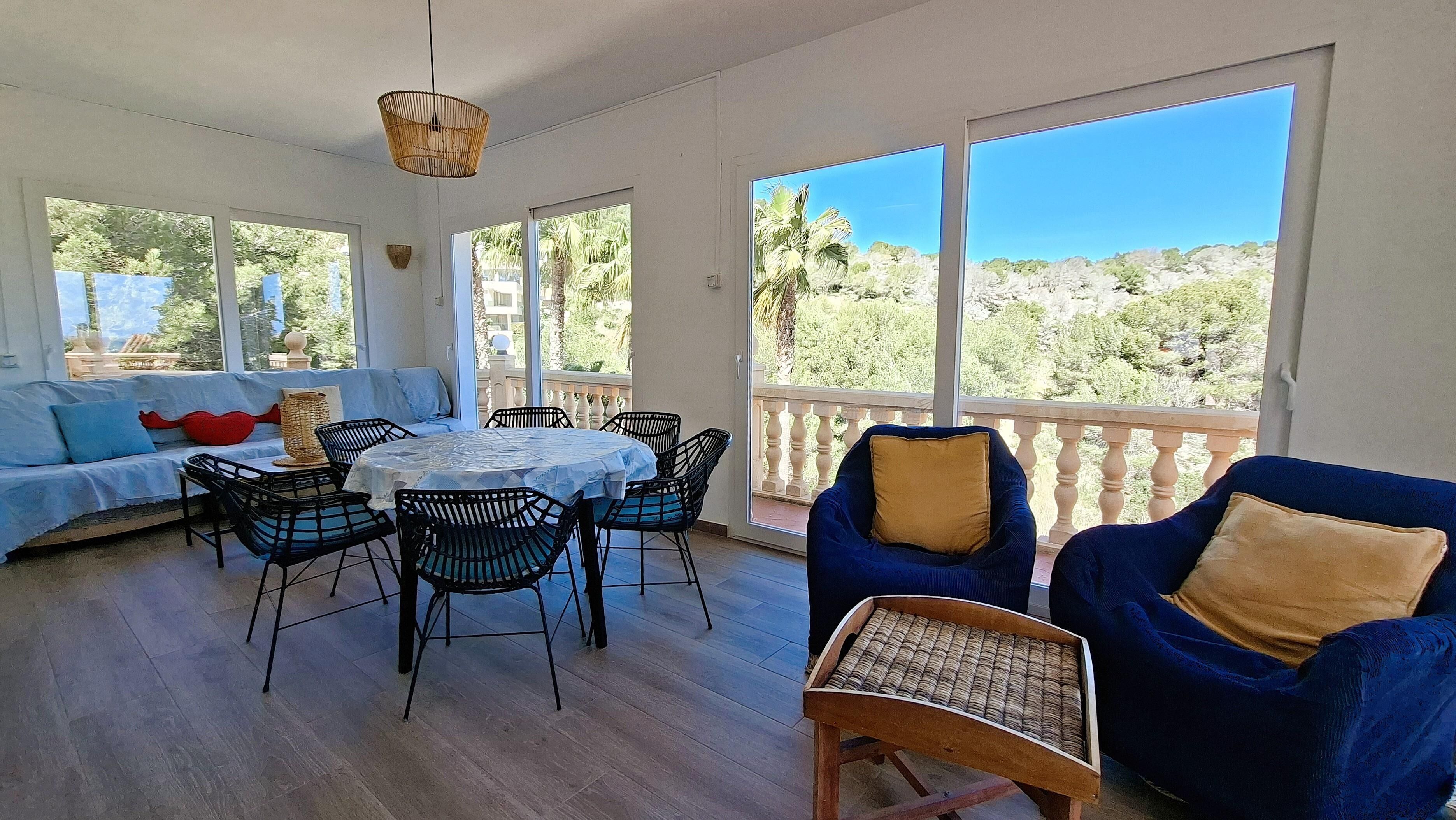 A sunlit room with a dining table, chairs, a sofa, and a balcony with a view of greenery.
