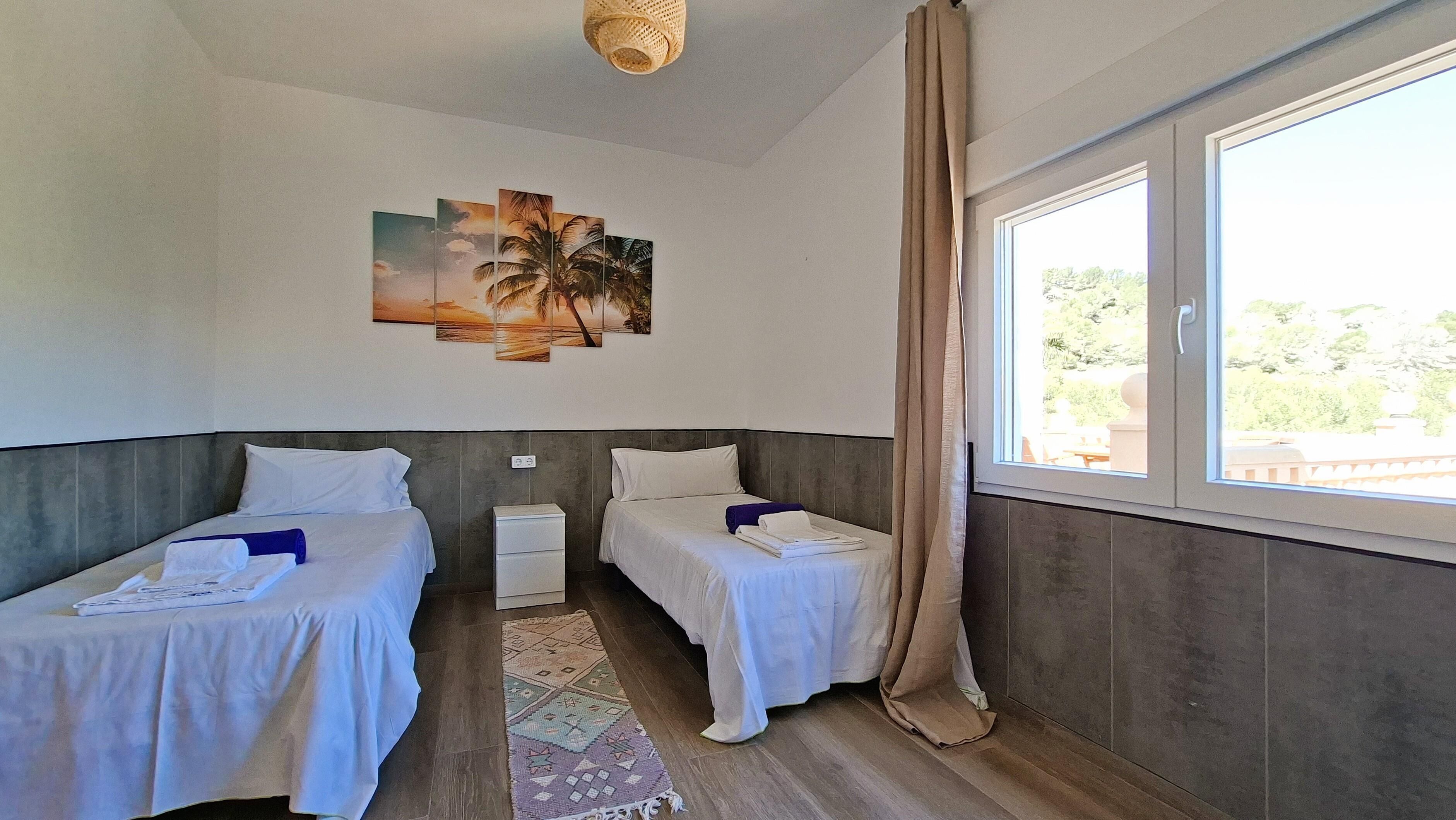 Ein Hotelzimmer mit zwei Betten, einem kleinen Nachttisch, einem an der Wand hängenden Kunstwerk, einem Fenster mit Vorhängen und Blick ins Grüne.