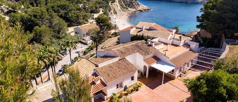 A coastal residential area with terracotta-tiled roofs, white buildings, and a beach in the background.