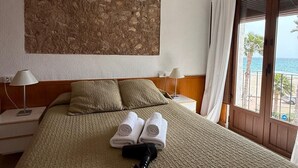 A hotel room with a bed, a ceiling fan, a large abstract painting, and a view of the sea through a door.