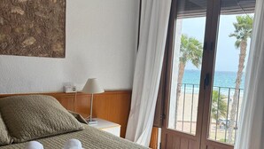 A hotel room with a bed, a nightstand, a lamp, a painting on the wall, and a balcony with a view of the sea and palm trees.