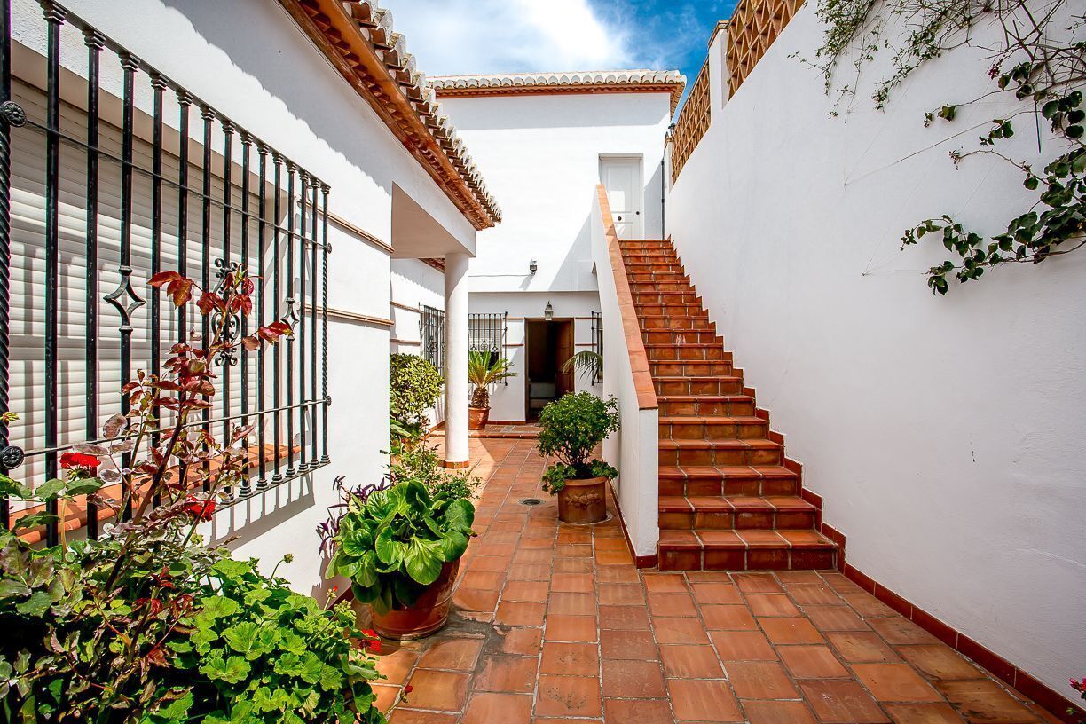 A narrow courtyard with a staircase leading to an upper level, surrounded by white walls and potted plants.