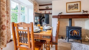 A dining area with a wooden table set for two, a fireplace, and a window with curtains. - Forsythia Cottage - sleeps 3 guests  in 2 bedrooms (Bourton-on-the-Water)