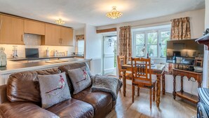 A cozy kitchen with wooden cabinets, a dining area with a wooden table and chairs, and a leather sofa with decorative pillows. - Forsythia Cottage - sleeps 3 guests  in 2 bedrooms (Bourton-on-the-Water)