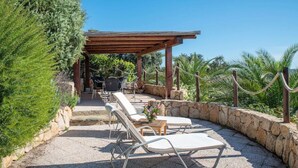 Une terrasse extérieure dotée de chaises longues et d’une pergola, entourée de murs en pierre et de verdure.