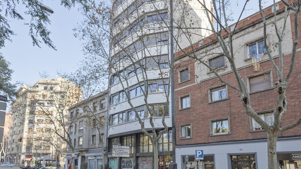 Un edificio de varios pisos con una mezcla de estilos arquitectónicos, incluyendo un edificio clásico con cúpula y una construcción moderna de ladrillo. Se observan indicaciones de dirección a diferentes lugares, y un cartel de estacionamiento es visible.