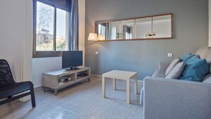 Una sala de estar moderna con un sofá gris, una mesita, un televisor sobre un mueble de madera y un ventanal con cortinas.