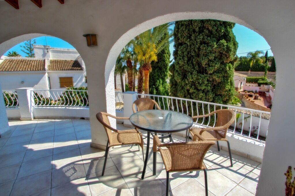 Un patio cubierto con una mesa de vidrio y sillas de mimbre, rodeado de muros blancos y una barandilla de metal.