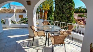 Une terrasse couverte avec une table en verre et des chaises en osier, entourée de murs blancs et d’une rambarde en métal.
