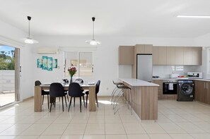 A modern kitchen with a central island, a dining area with chairs, and a view through a glass door.